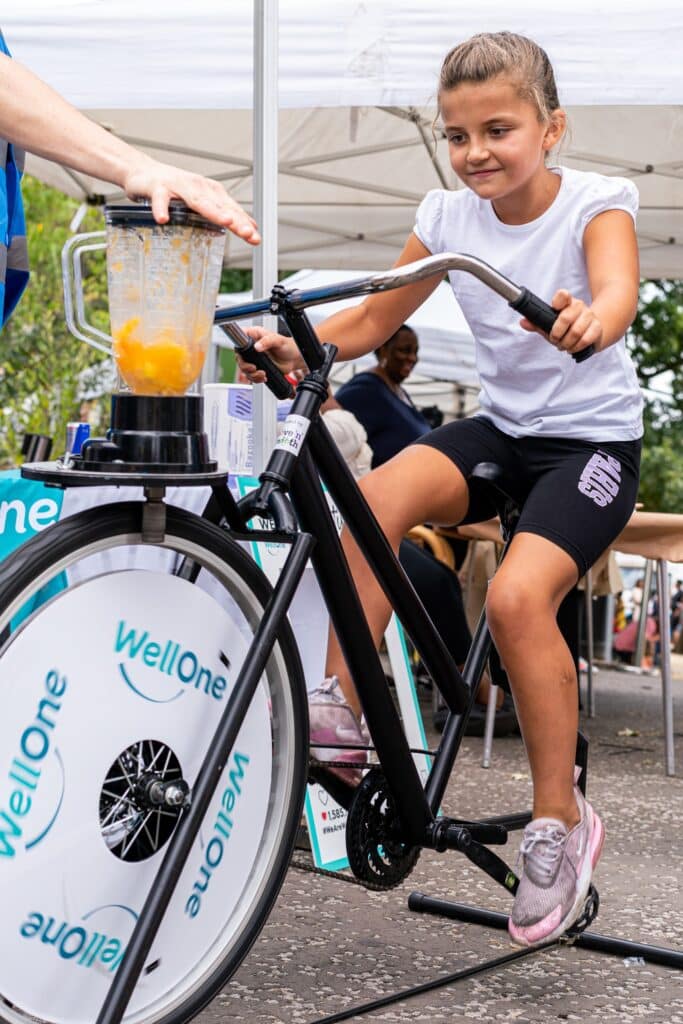 A girl is pedalling to make a fruit smoothie on the Well One smoothie bike at the Summer of Wellbeing festival.
