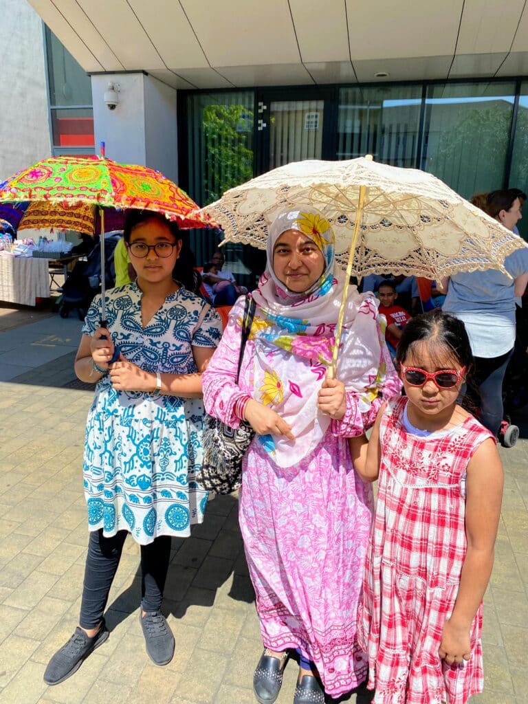 A mother and her two daughters attending the Summer of Wellbeing Festival in Poplar, London. They are dressed in colourful patterned clothes and using traditional Bengali parasols to shade from the sun.