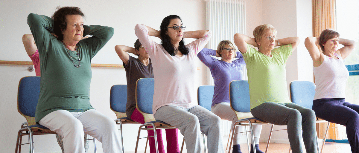 People at a seated exercise class, gently stretching with their hands behind their heads.